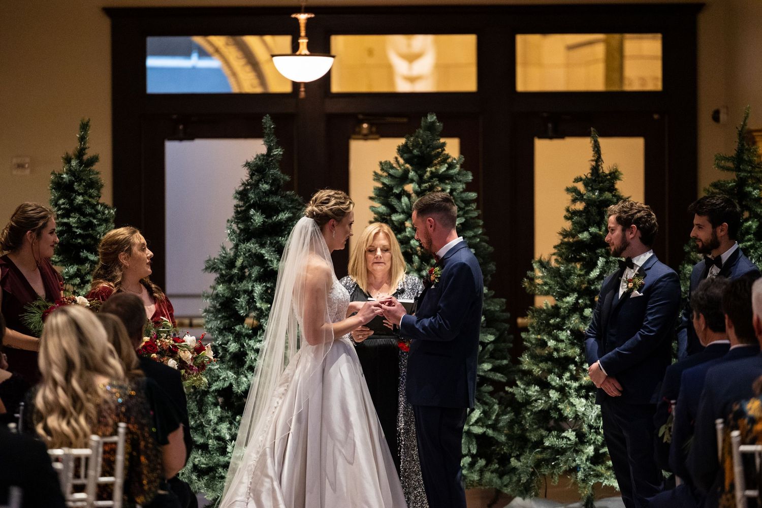 Wedding ceremony in progress with decorated Christmas trees flanking the aisle.