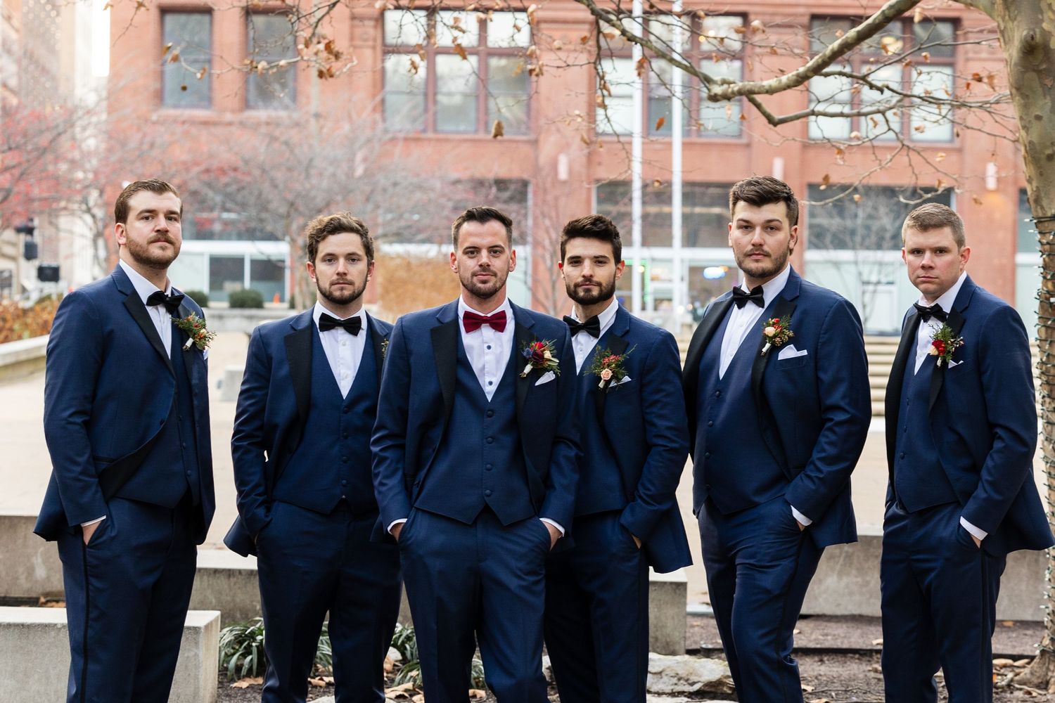 Groomsmen in navy suits pose together outdoors against brick building backdrop.