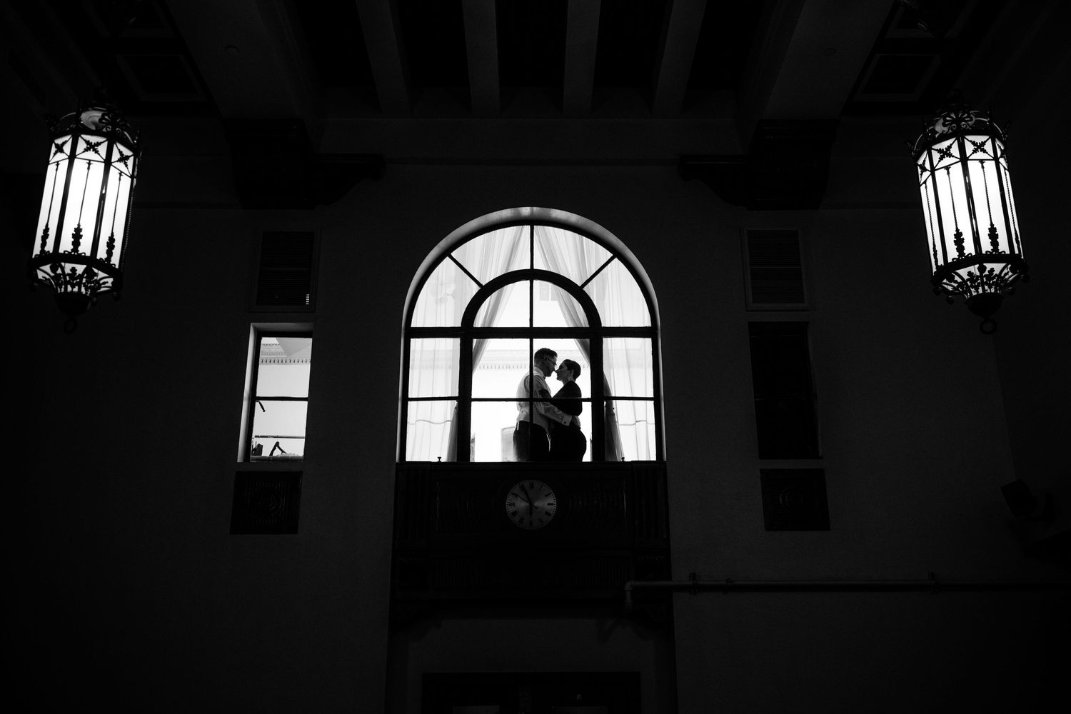 Silhouetted figures against arched window with hanging lanterns creating dramatic wedding venue atmosphere.
