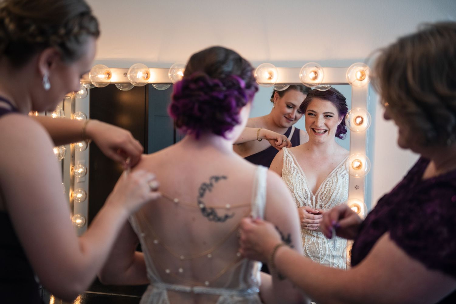 Wedding party helping with final dress preparations in front of a vanity mirror with marquee lights.