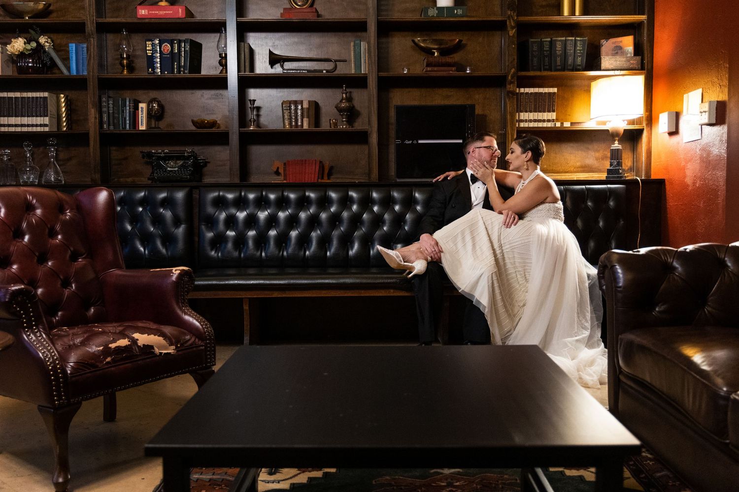 Couple in wedding attire sits on leather couch in library setting.