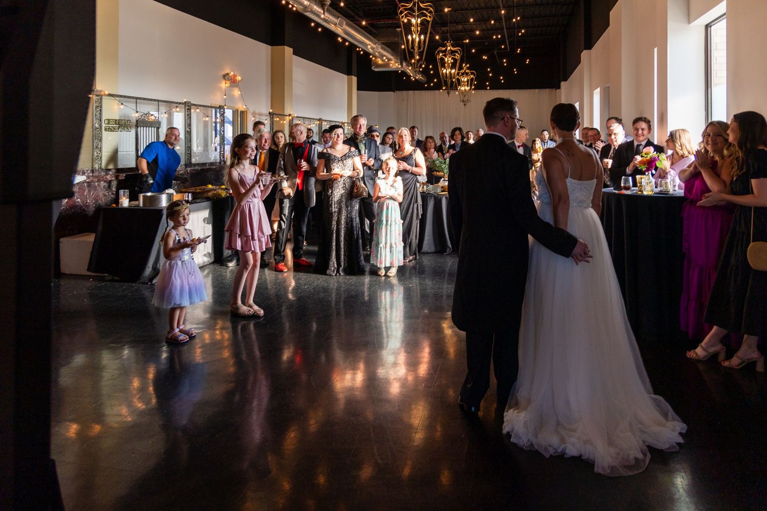 Wedding reception dance floor with guests gathered around couple in elegant venue with string lights.