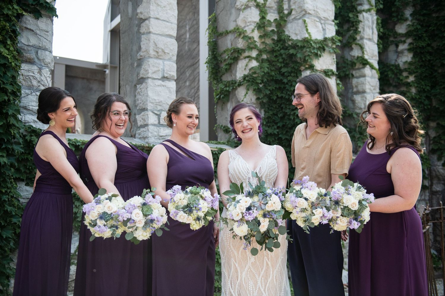 Bridesmaids in purple dresses hold white and purple bouquets while standing with bride.