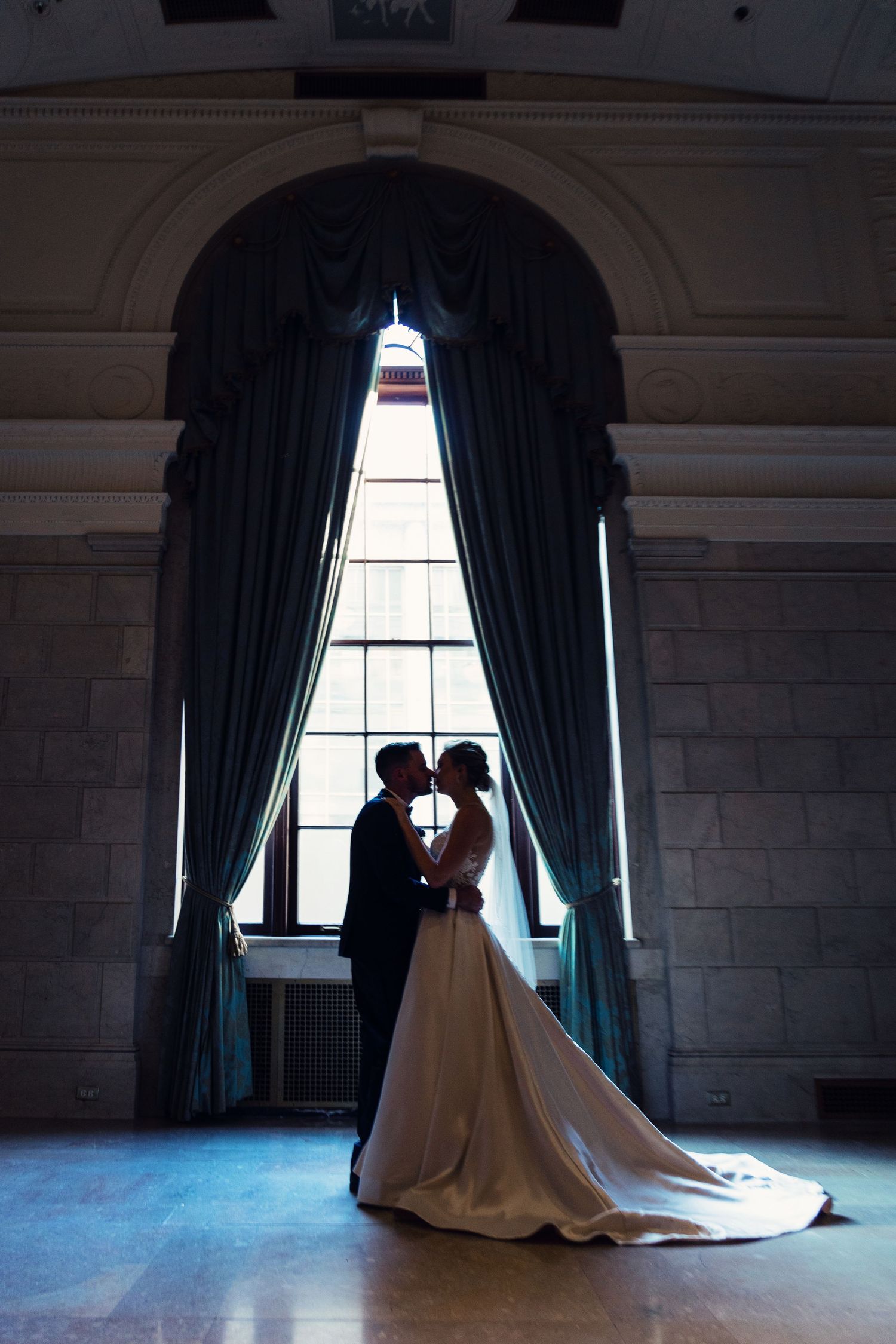 Silhouetted wedding couple in dramatic window light with long curtains and arched architecture.