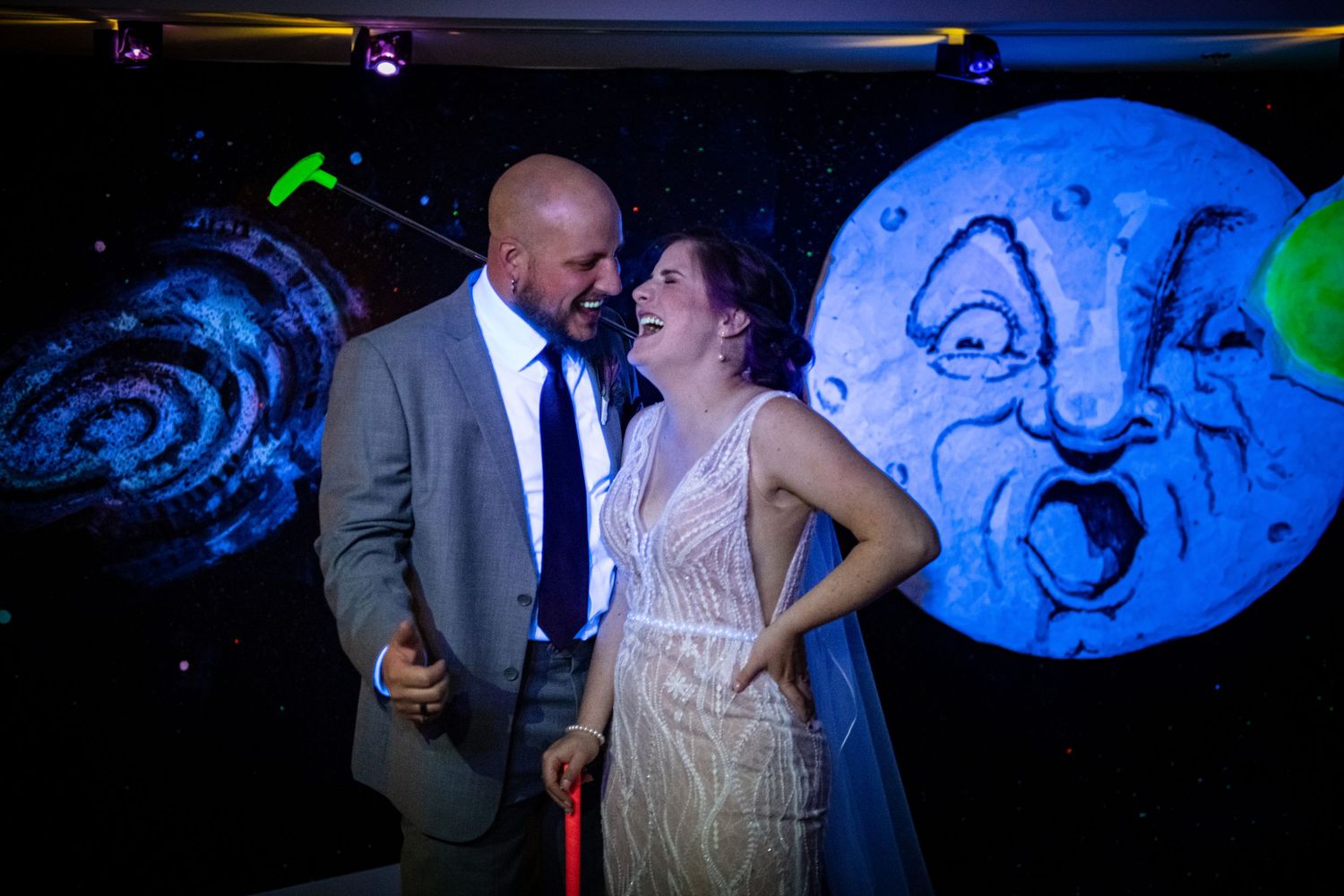 A couple poses in front of a whimsical moon backdrop with colorful lighting.