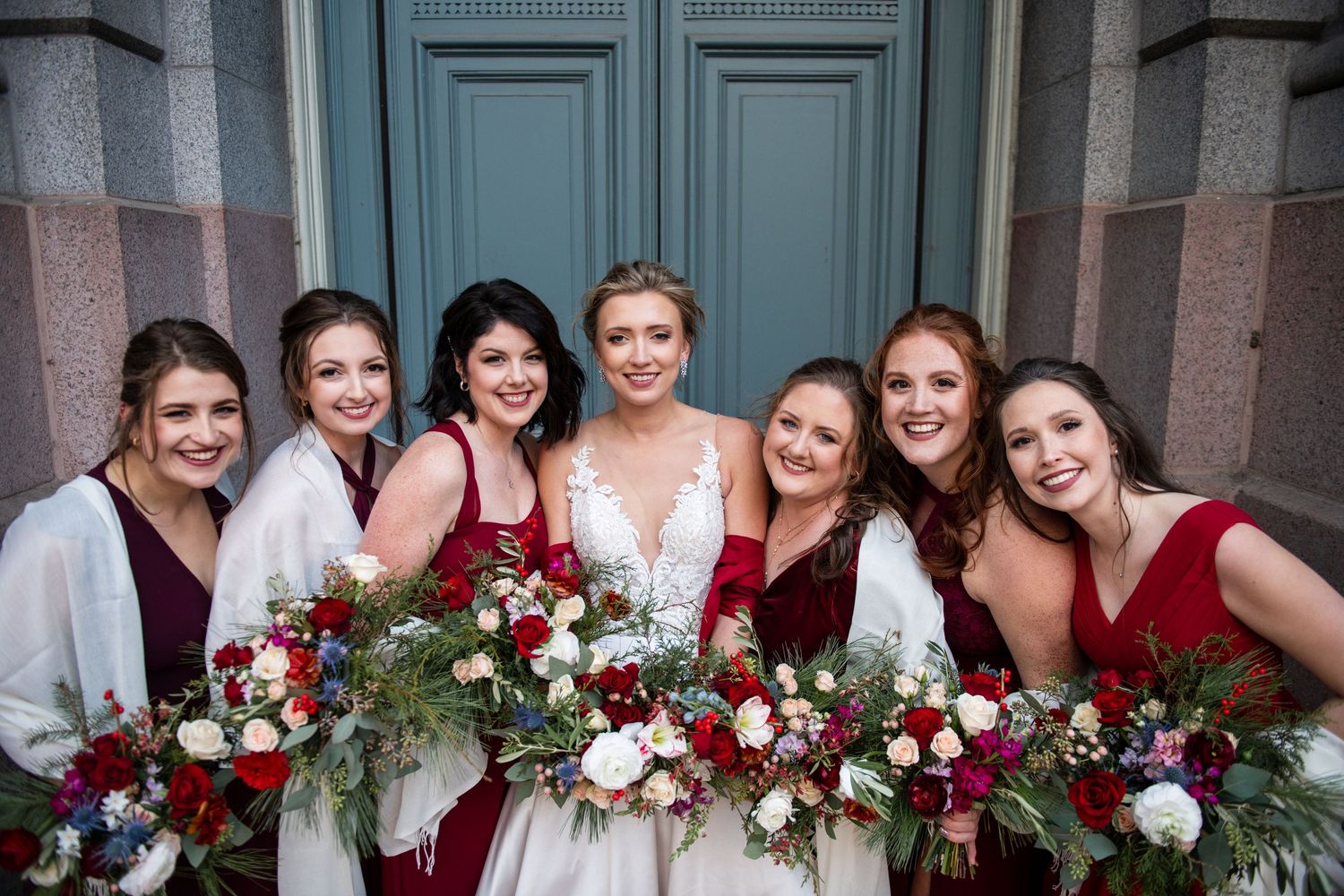 Bridesmaids with burgundy dresses gather around bride holding winter floral arrangements.