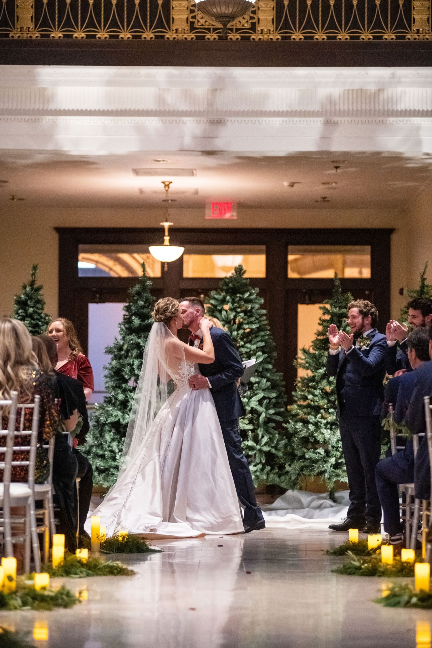 Couple sharing their first kiss during winter wedding ceremony surrounded by Christmas trees.
