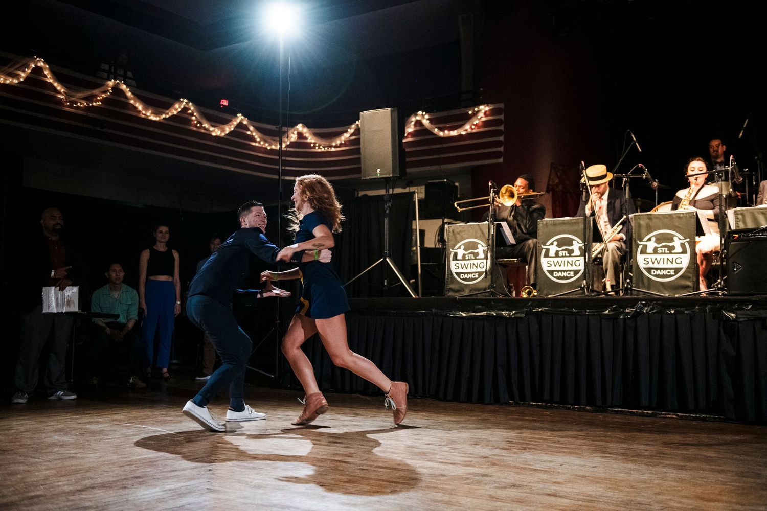 Dancers perform energetic swing dance moves in front of live band on elevated stage.