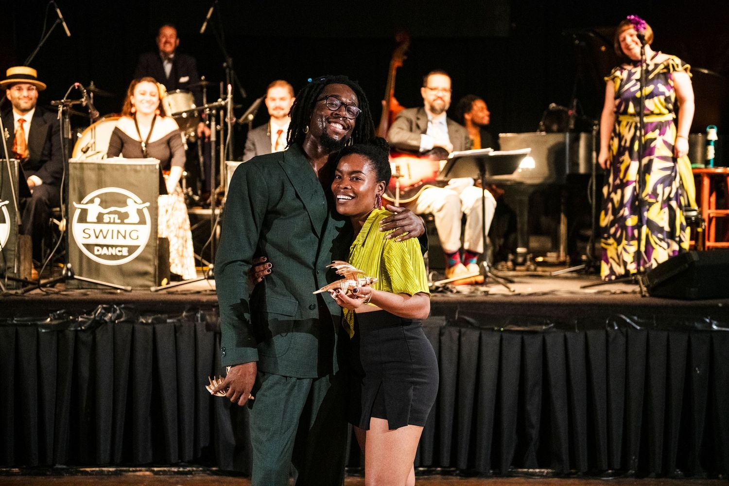 Two people stand on stage receiving awards with band members visible in the background.