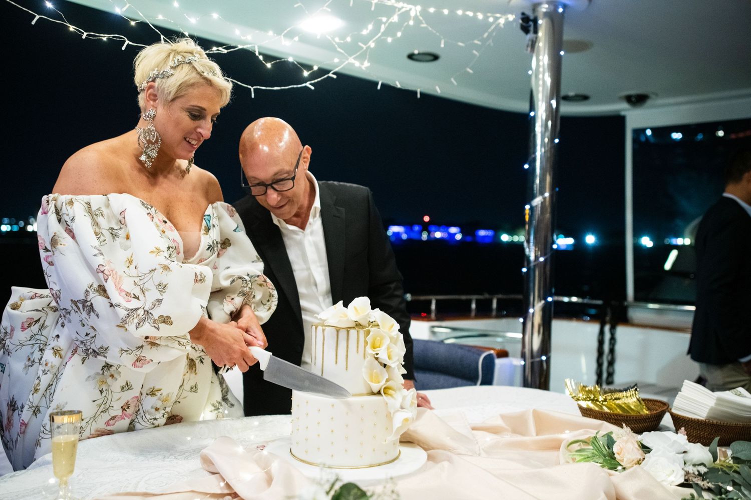A couple sharing a moment while cutting their white and gold wedding cake at an evening reception.