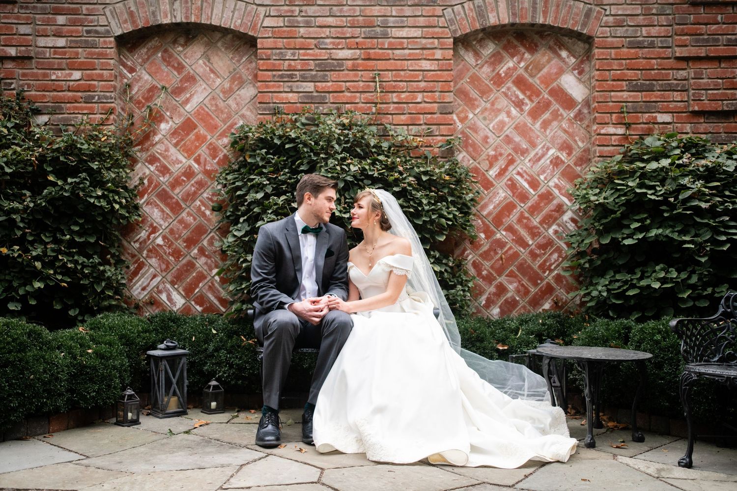 Wedding couple seated on bench against brick wall with decorative arched insets.