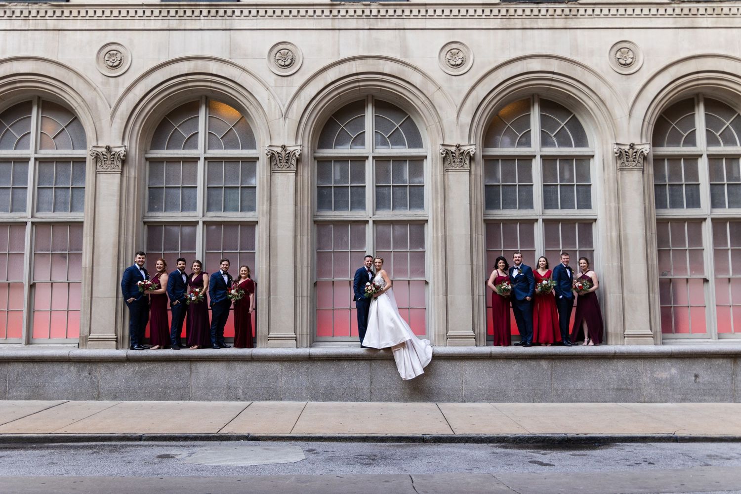 Wedding party poses in arched windows of historic building facade.