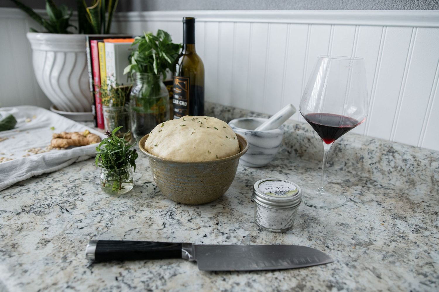 Fresh bread dough rising in a ceramic bowl with herbs, wine and cooking utensils on marble countertop.