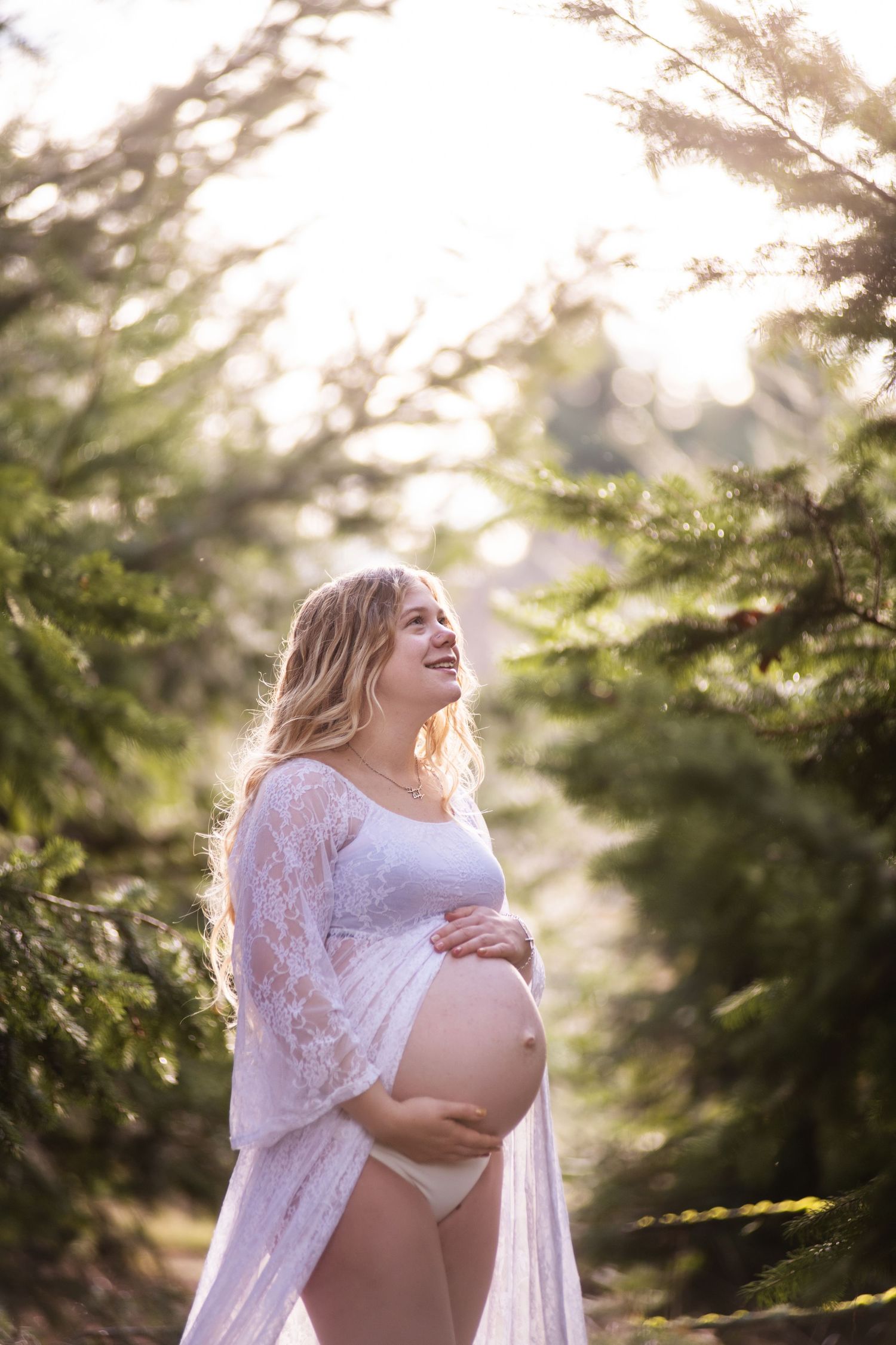 A pregnant figure in a white lace gown poses ethereally among evergreen trees during a sunlit maternity photoshoot.