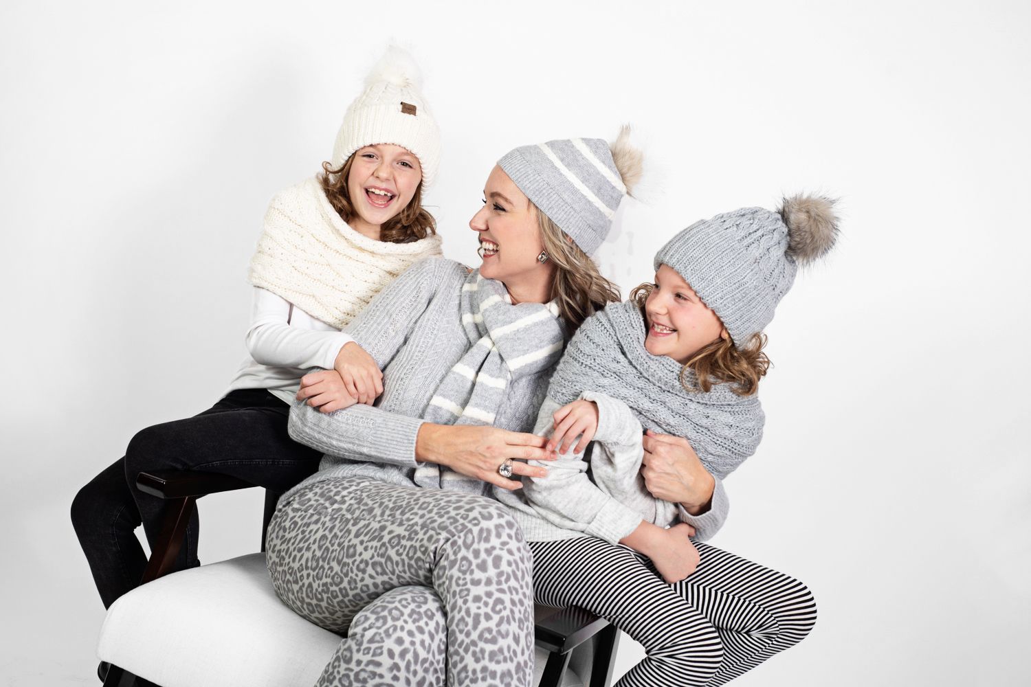 Three people in cozy winter hats and sweaters laughing together on white furniture.