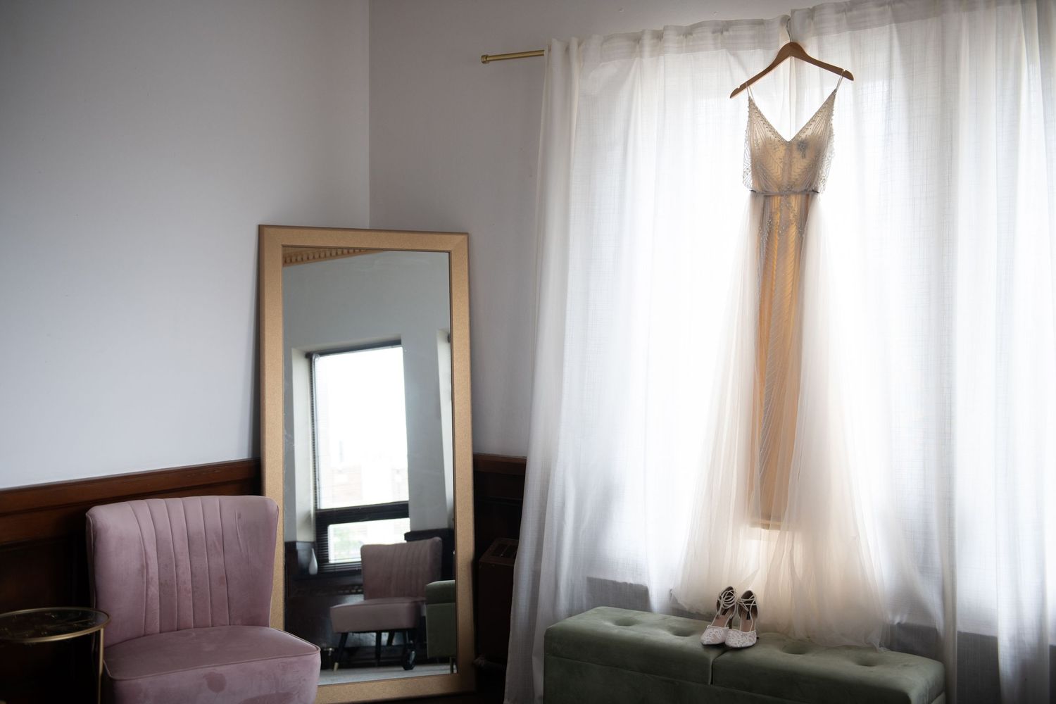 Wedding dress hanging by window with full-length mirror and chair in getting ready room.
