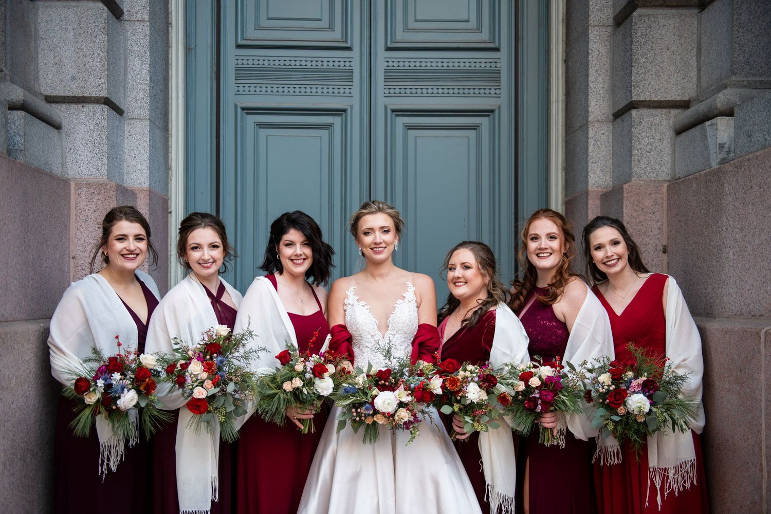 Bridal party in burgundy dresses and white shawls pose with winter floral bouquets by blue doors.