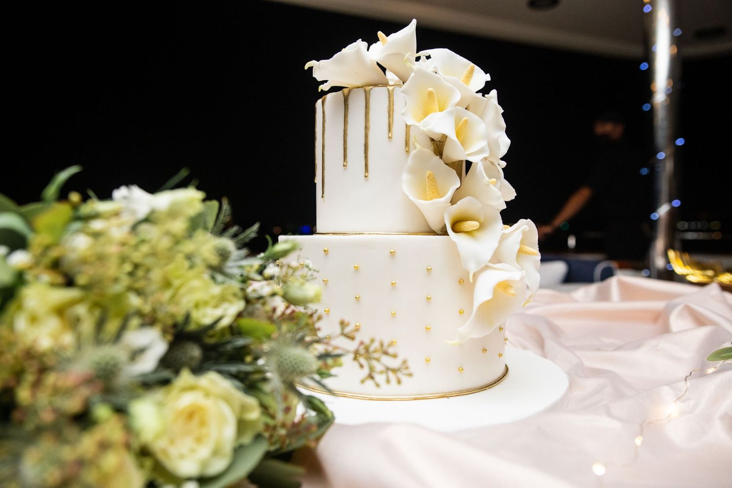 Elegant white wedding cake with gold accents and cascading floral decorations displayed on a table with flower arrangements.