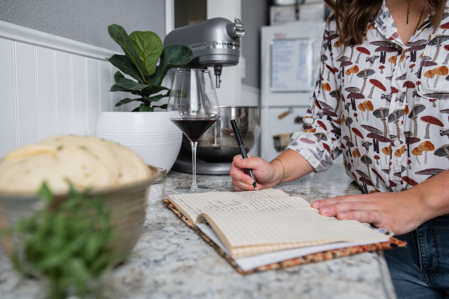 Writing in a notebook beside a glass of wine on a marble countertop with fresh herbs nearby.