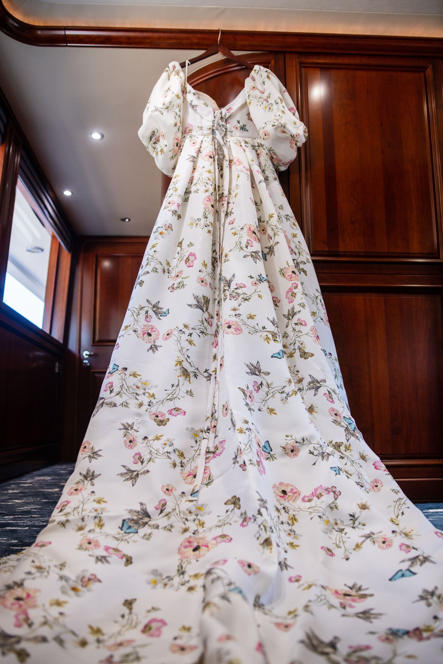 Full-length view of romantic floral print wedding gown with puff sleeves hanging in yacht cabin.