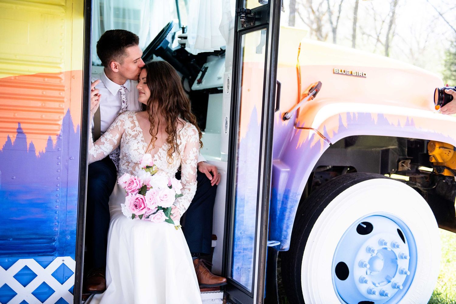 Newlyweds share a playful moment by a colorful bus during their wedding celebration.