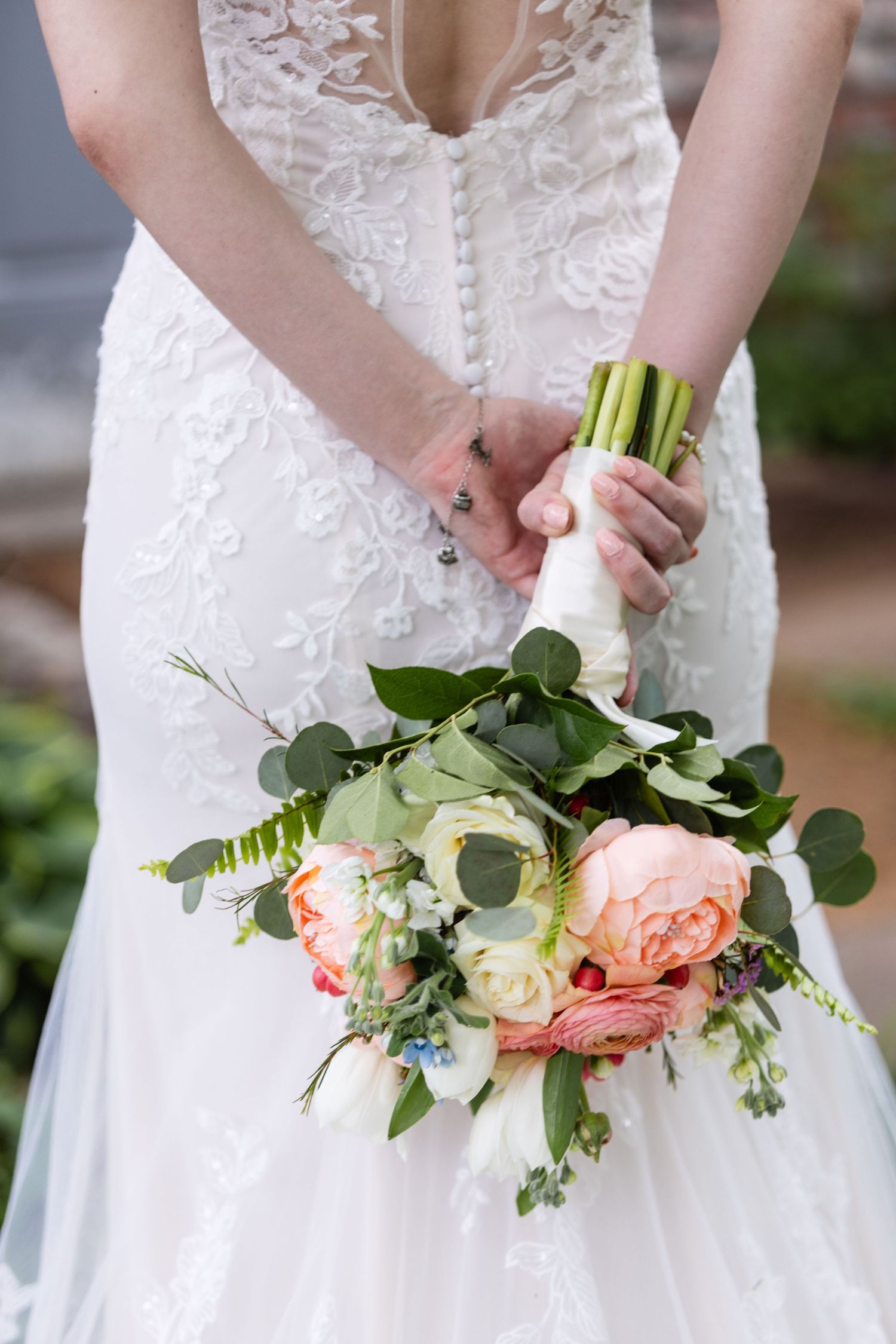 Close-up of bridal bouquet with peach roses and eucalyptus held against white lace dress.