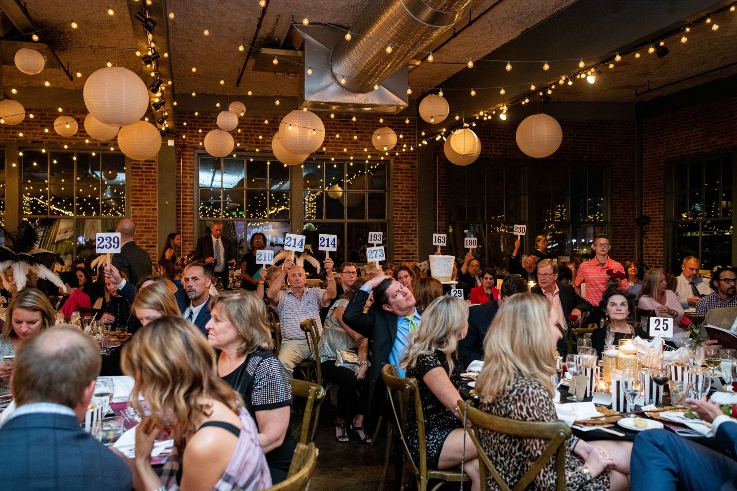 Lively auction dinner event with round tables and decorative lighting in an industrial-style venue.