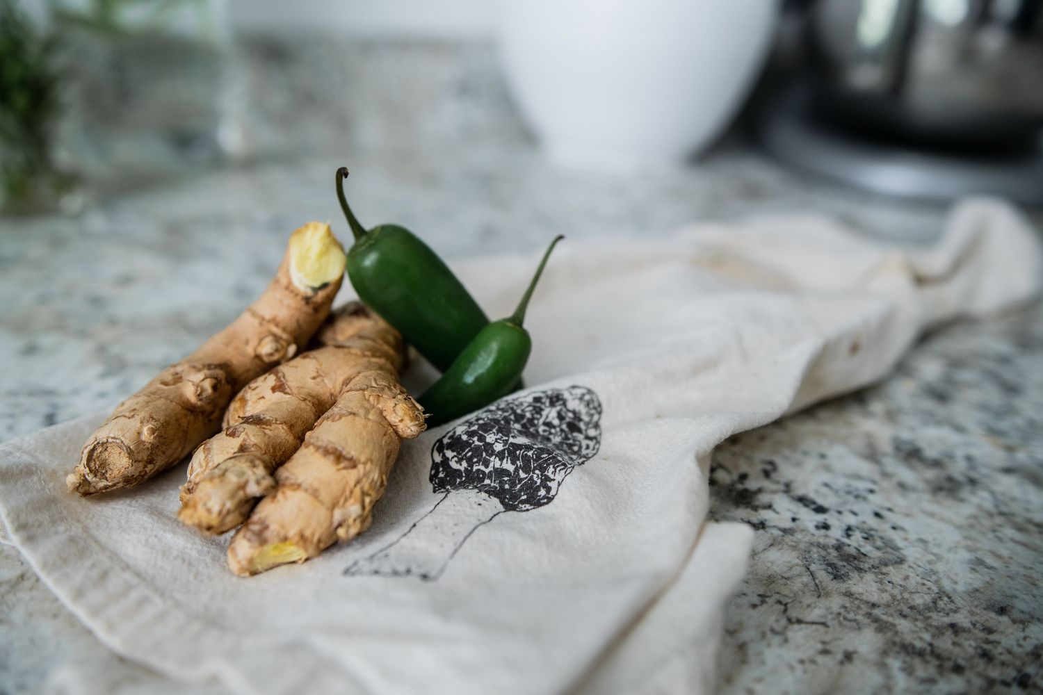 Fresh ginger root and green peppers arranged on a white cloth napkin on marble counter.