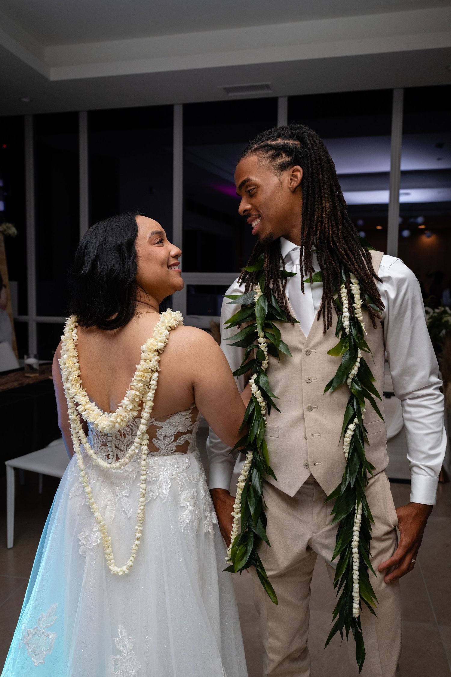 A couple shares a romantic moment at a wedding reception wearing traditional Hawaiian leis and floral decorations.