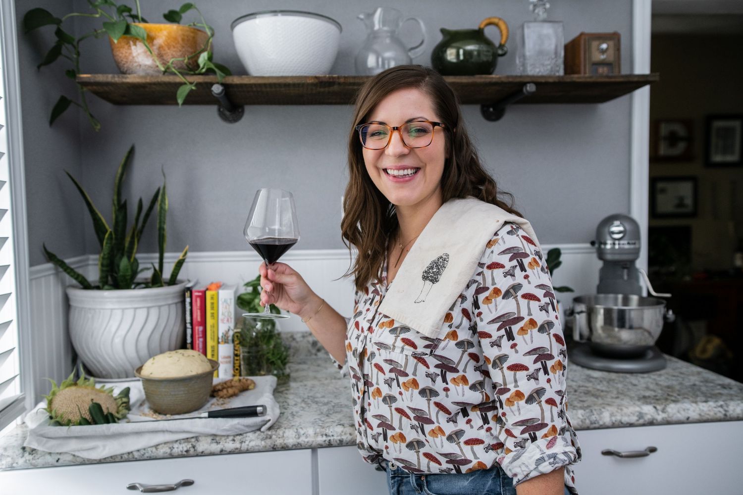 A cook raises a glass of wine while standing in a kitchen with decorative shelving.