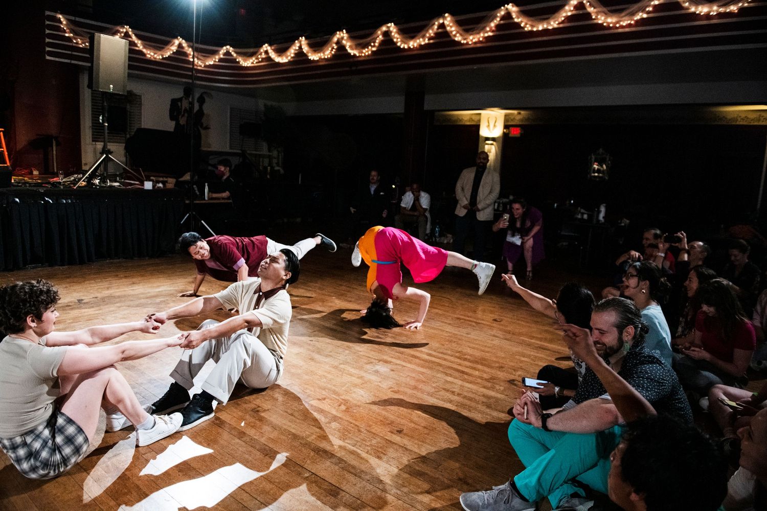 Break dancers performing acrobatic moves on wooden dance floor while audience watches.