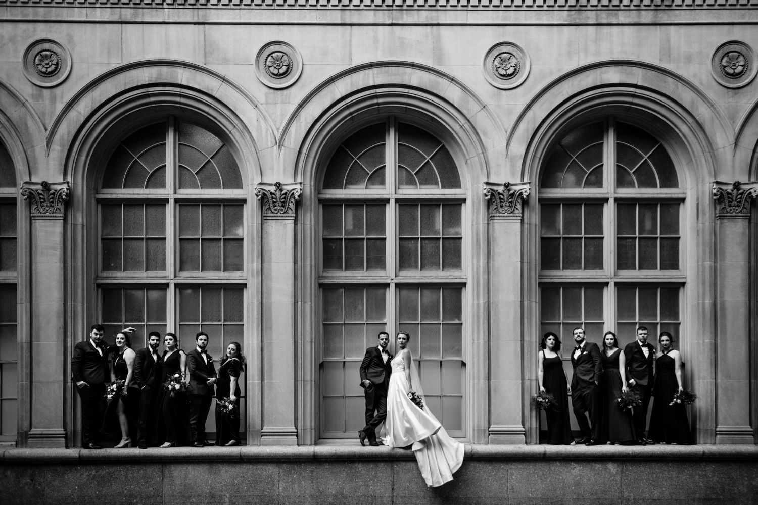 Black and white wedding portrait with bridal party arranged along classical arched windows.