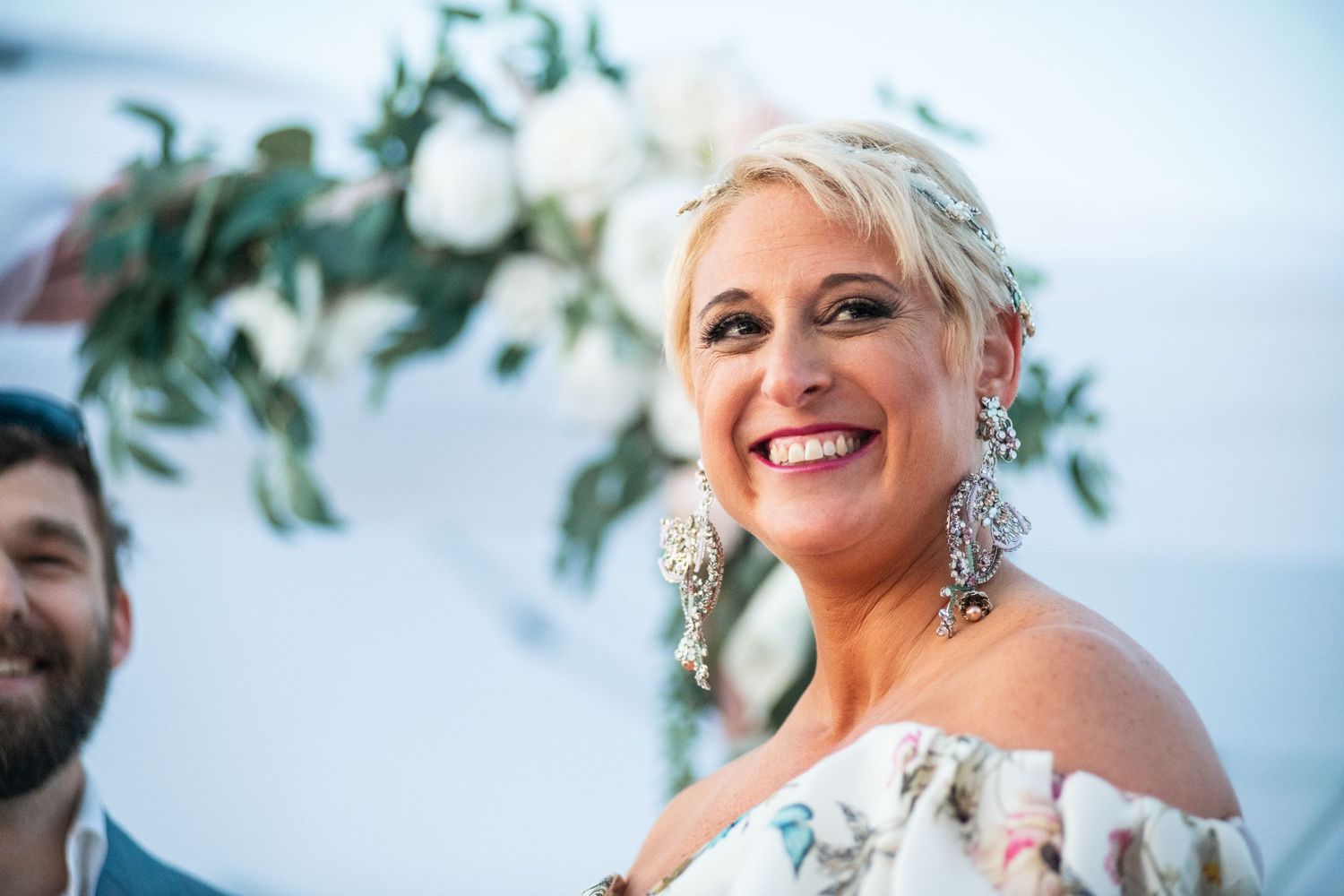 A bride in an off-shoulder floral dress smiles joyfully during a waterfront wedding ceremony.