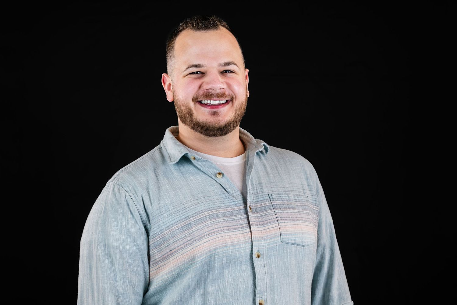 Professional headshot portrait of someone wearing a light chambray button-up shirt against a black background.
