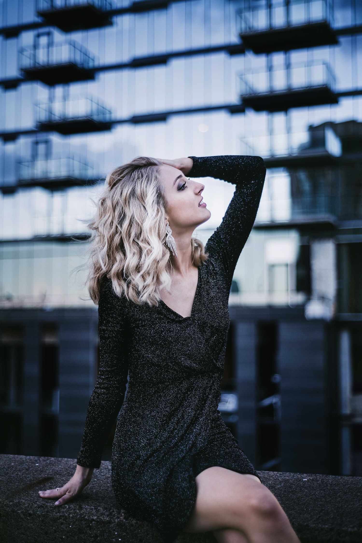 Artistic urban fashion portrait with sparkly black dress against modern building facade at dusk.
