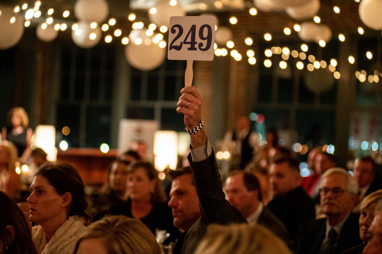 A person holds up auction paddle number 249 during an evening event with twinkling lights in the background.