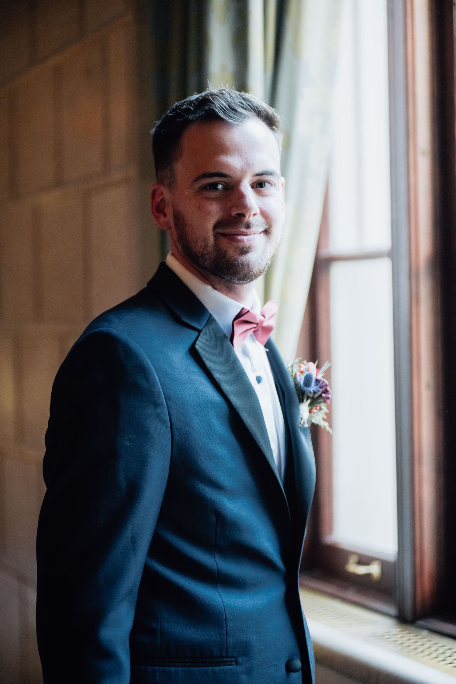 Groom in navy suit with pink boutonniere poses by window in elegant interior.