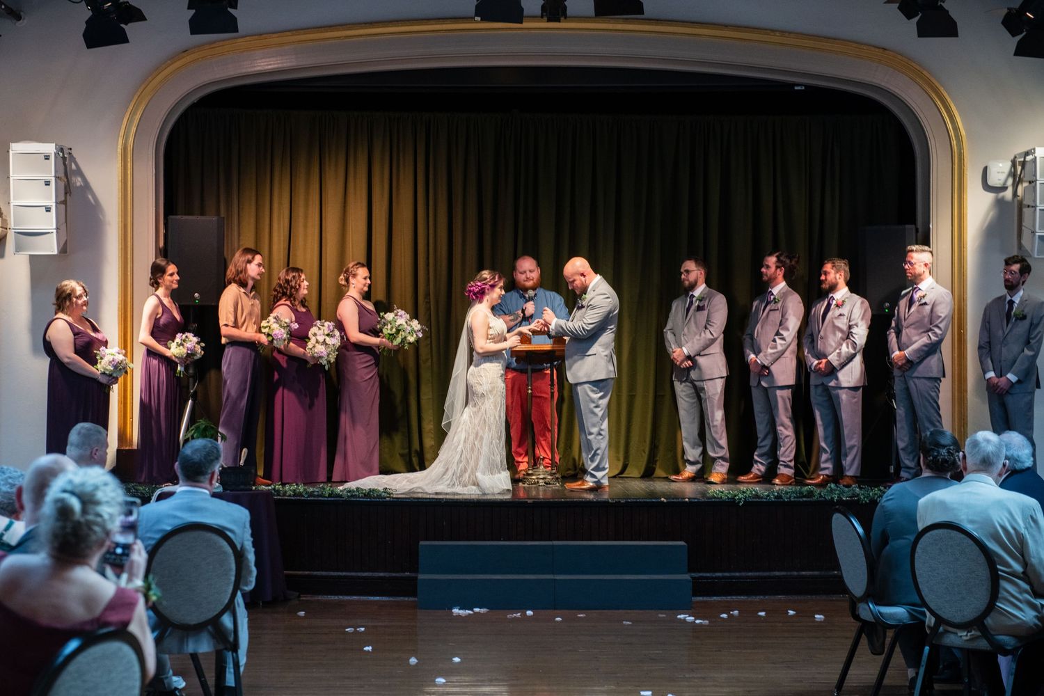 Wedding ceremony on stage with bridal party and audience in a vintage theater setting.