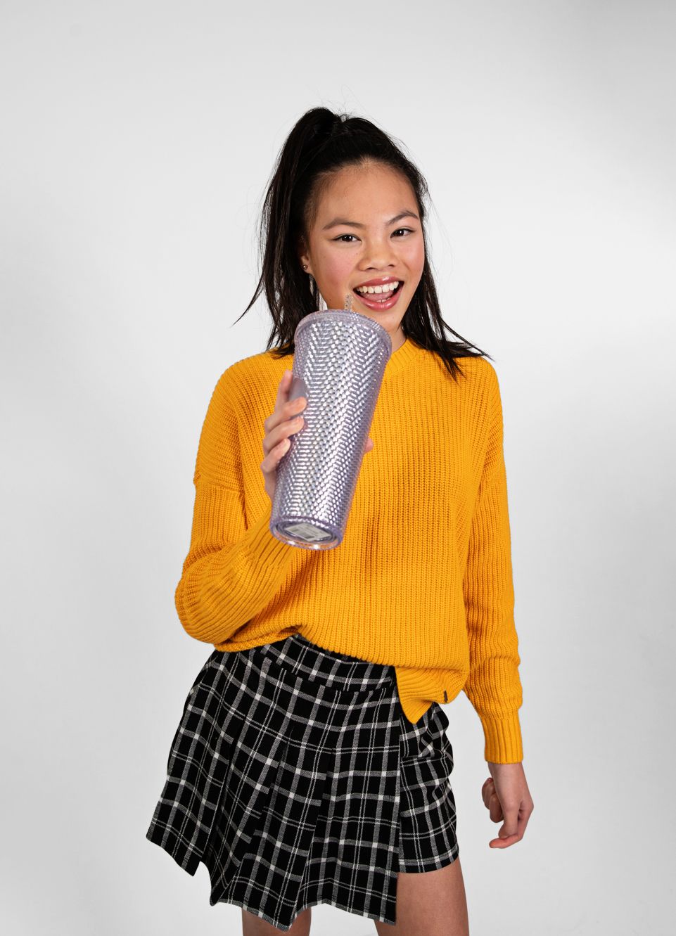 Young woman in yellow sweater and plaid skirt laughing while holding silver tumbler.