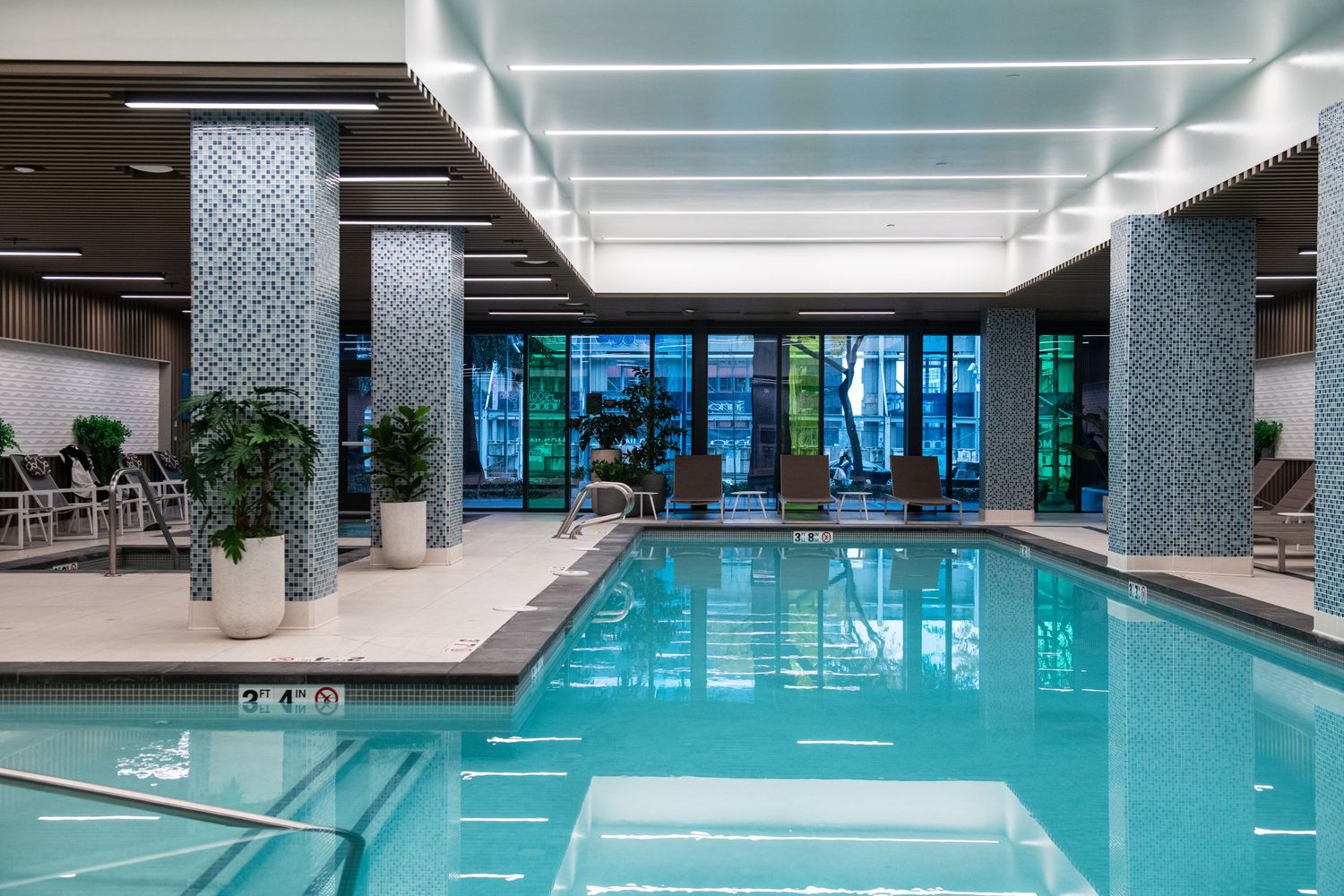 Modern indoor swimming pool with large windows and lounge chairs in luxurious apartment complex.
