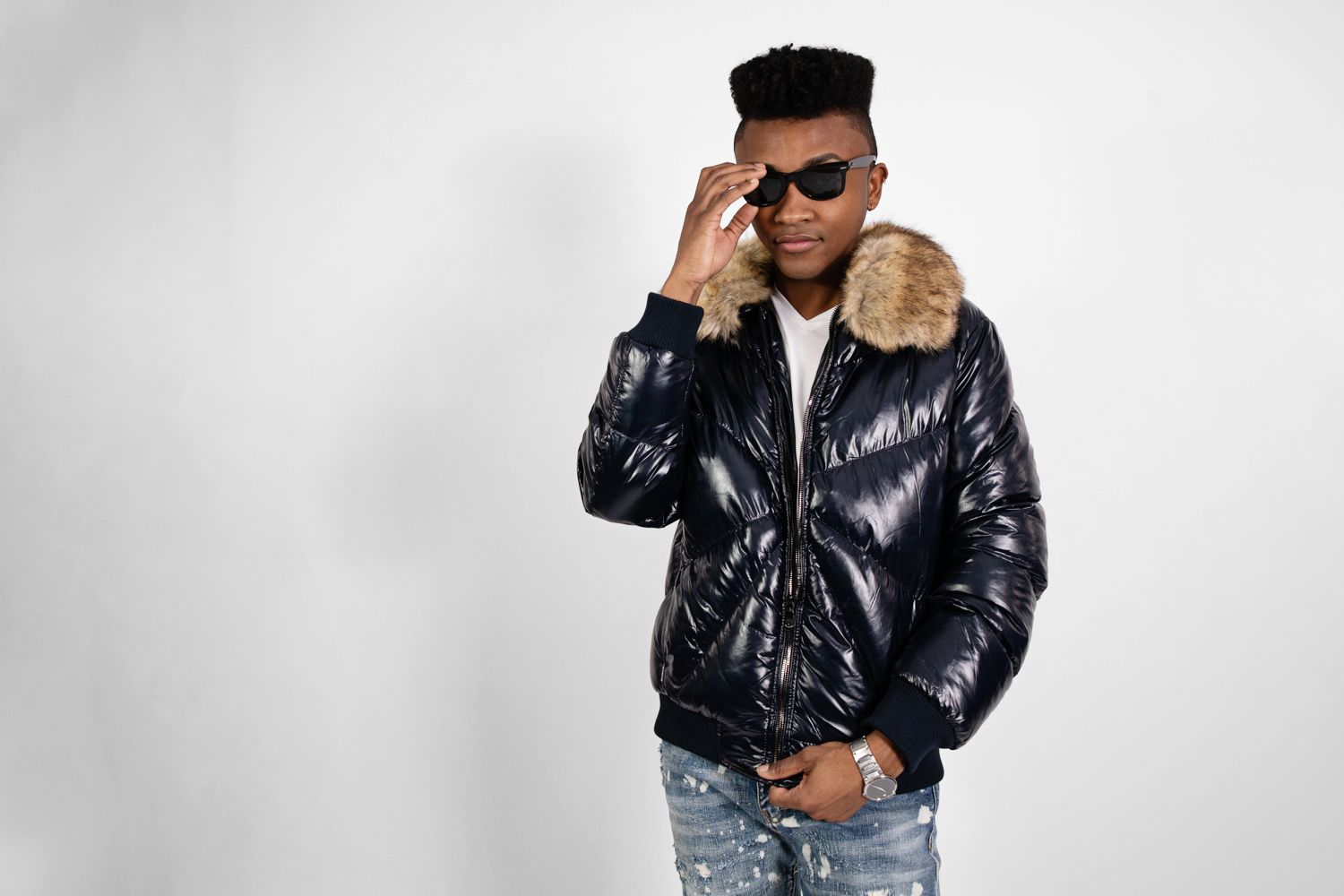 Fashion model adjusts sunglasses while wearing shiny puffer coat with fur collar.