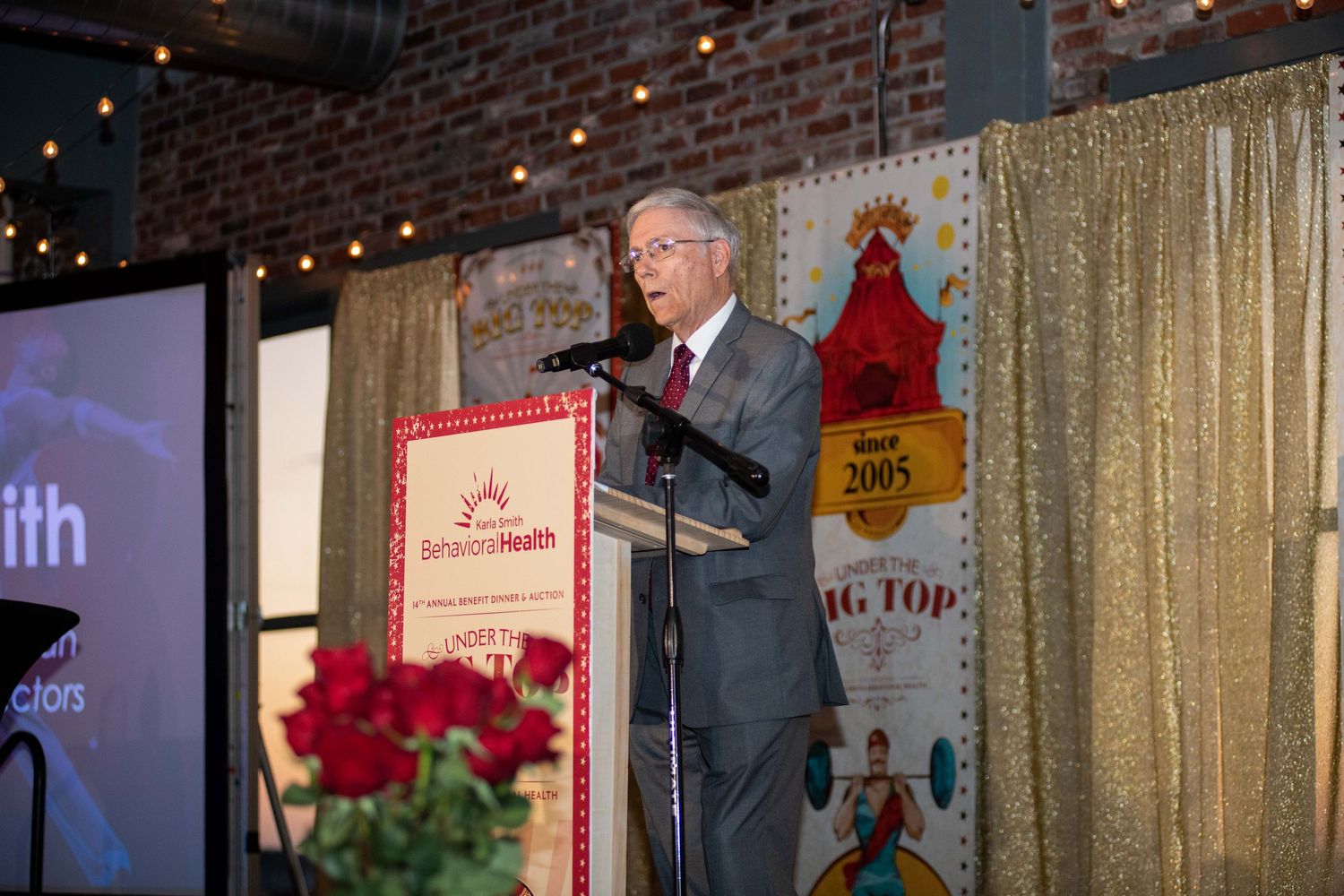 Speaker at podium presenting during formal event with red roses and glittering gold backdrop.