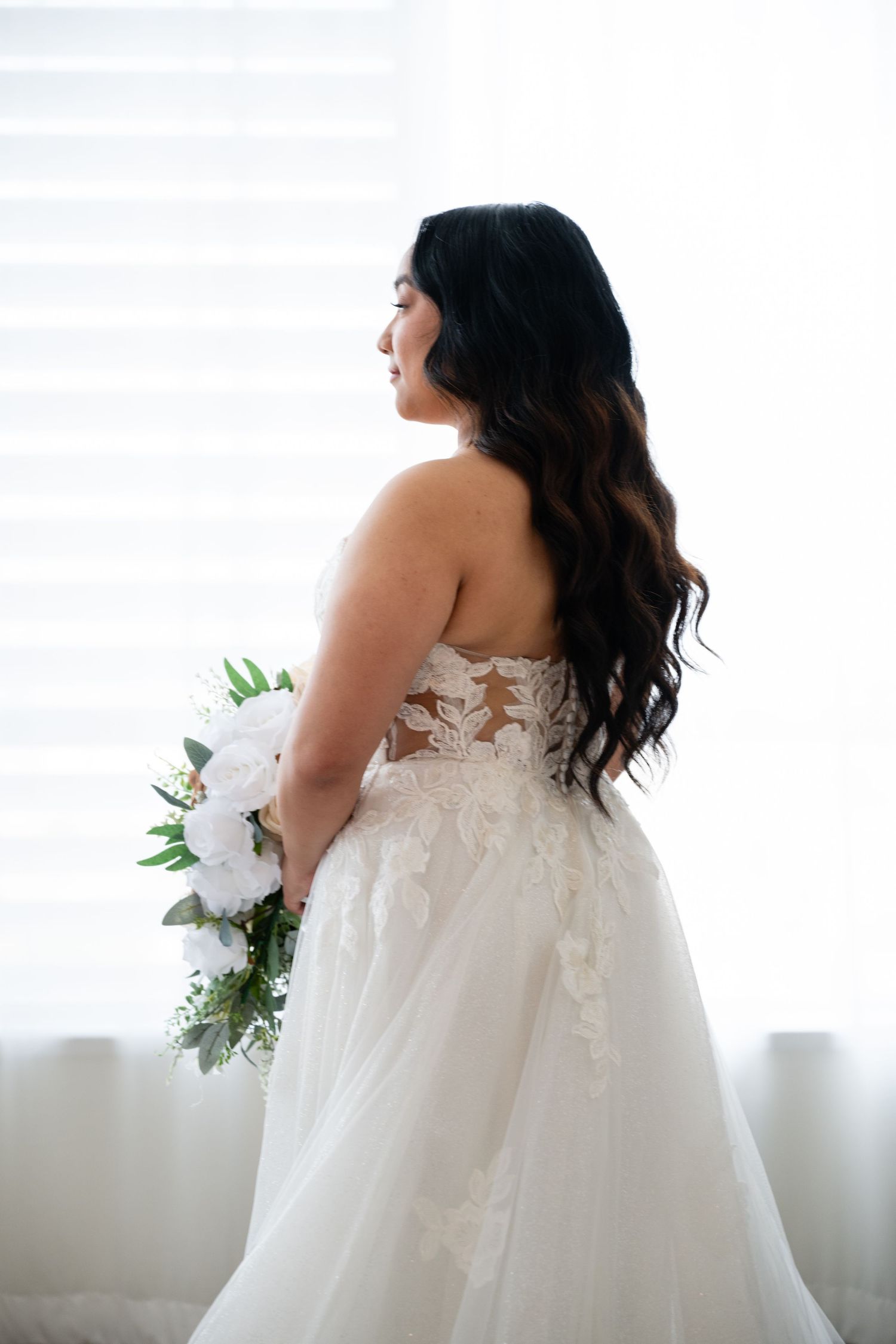 A bride in an elegant white lace wedding dress holds a floral bouquet in a series of poses near a window.
