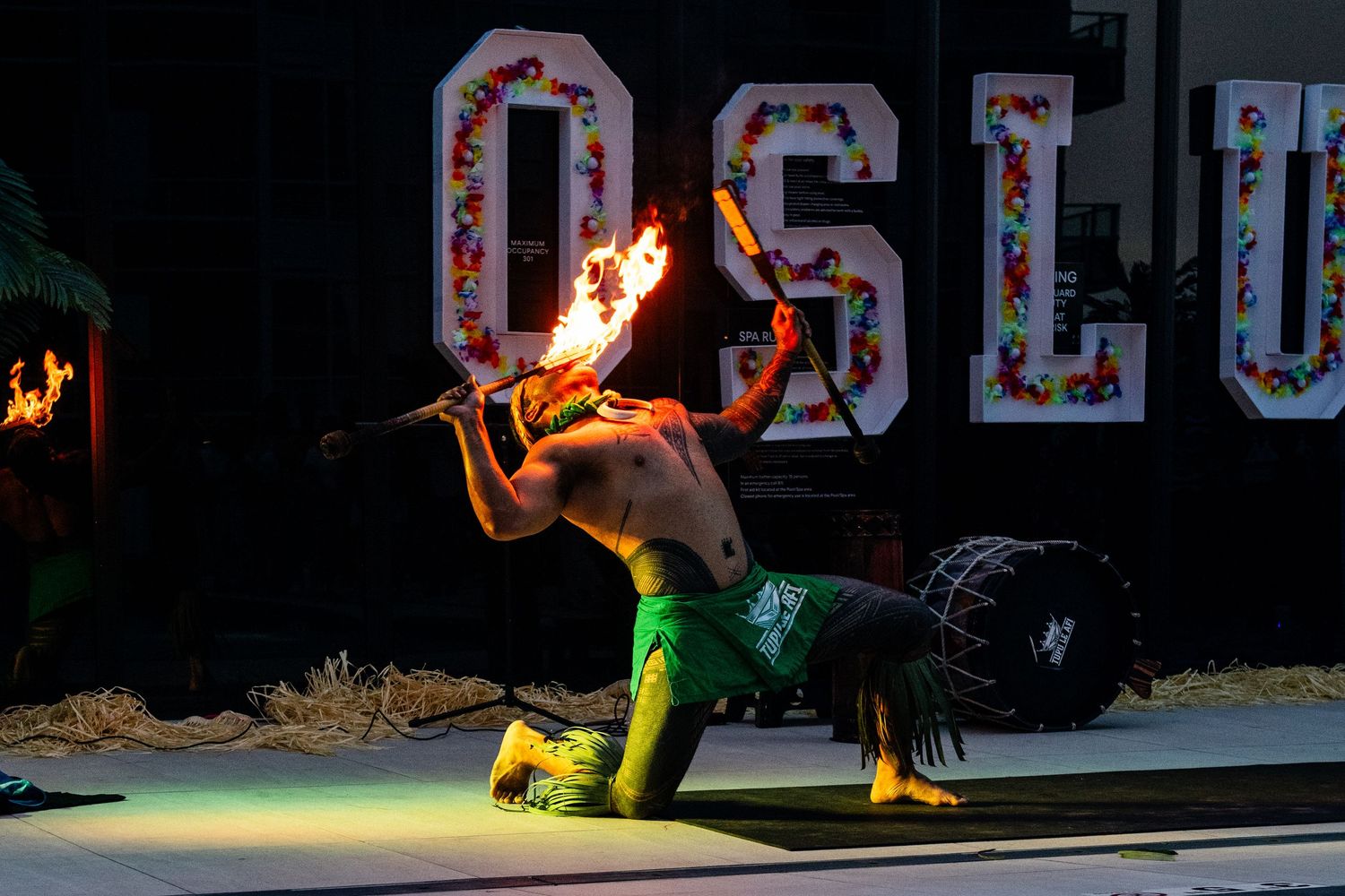 Dramatic night performance of fire dancer in green outfit against illuminated sign backdrop.