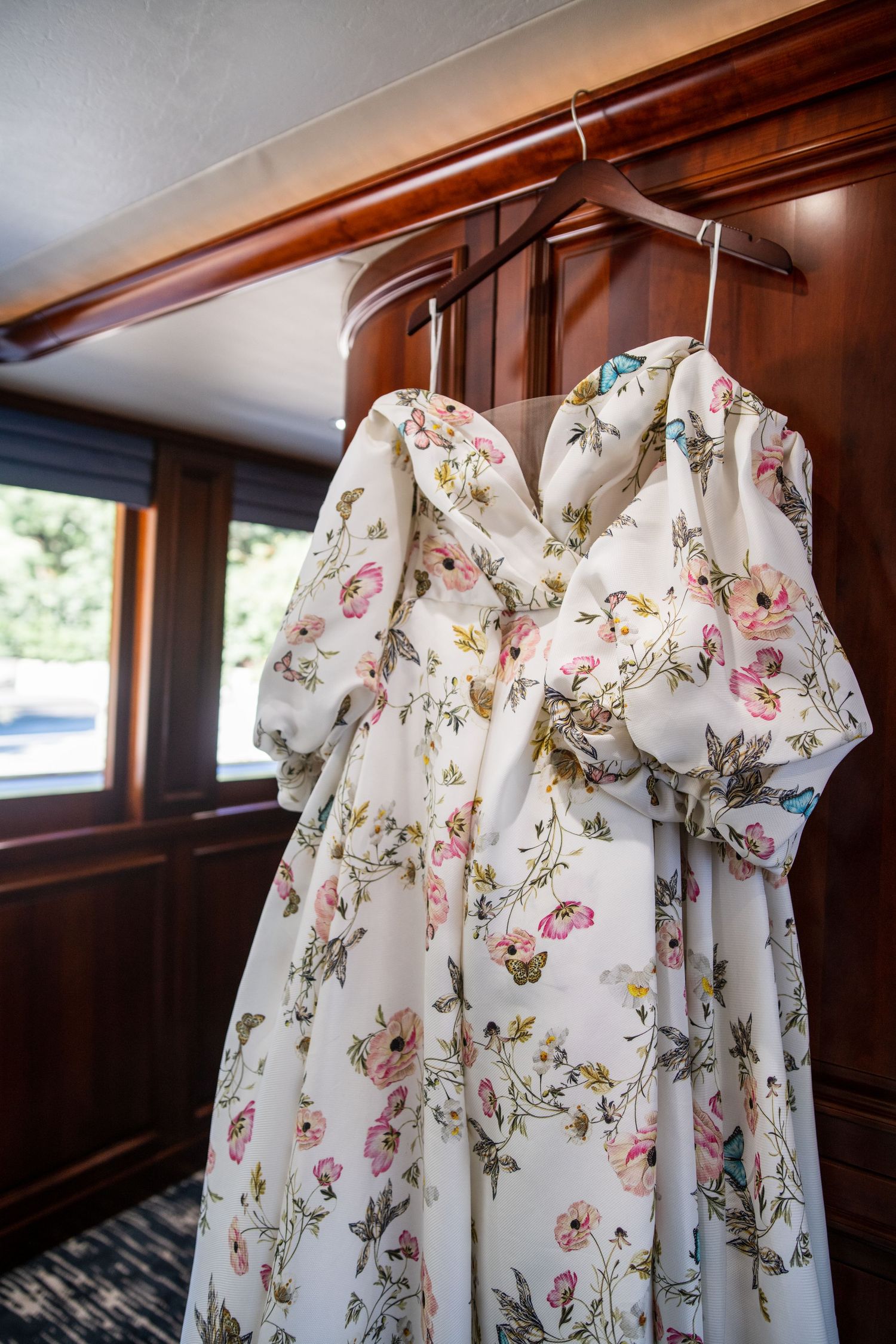 Floral print wedding dress hanging in wood-paneled room with natural light.