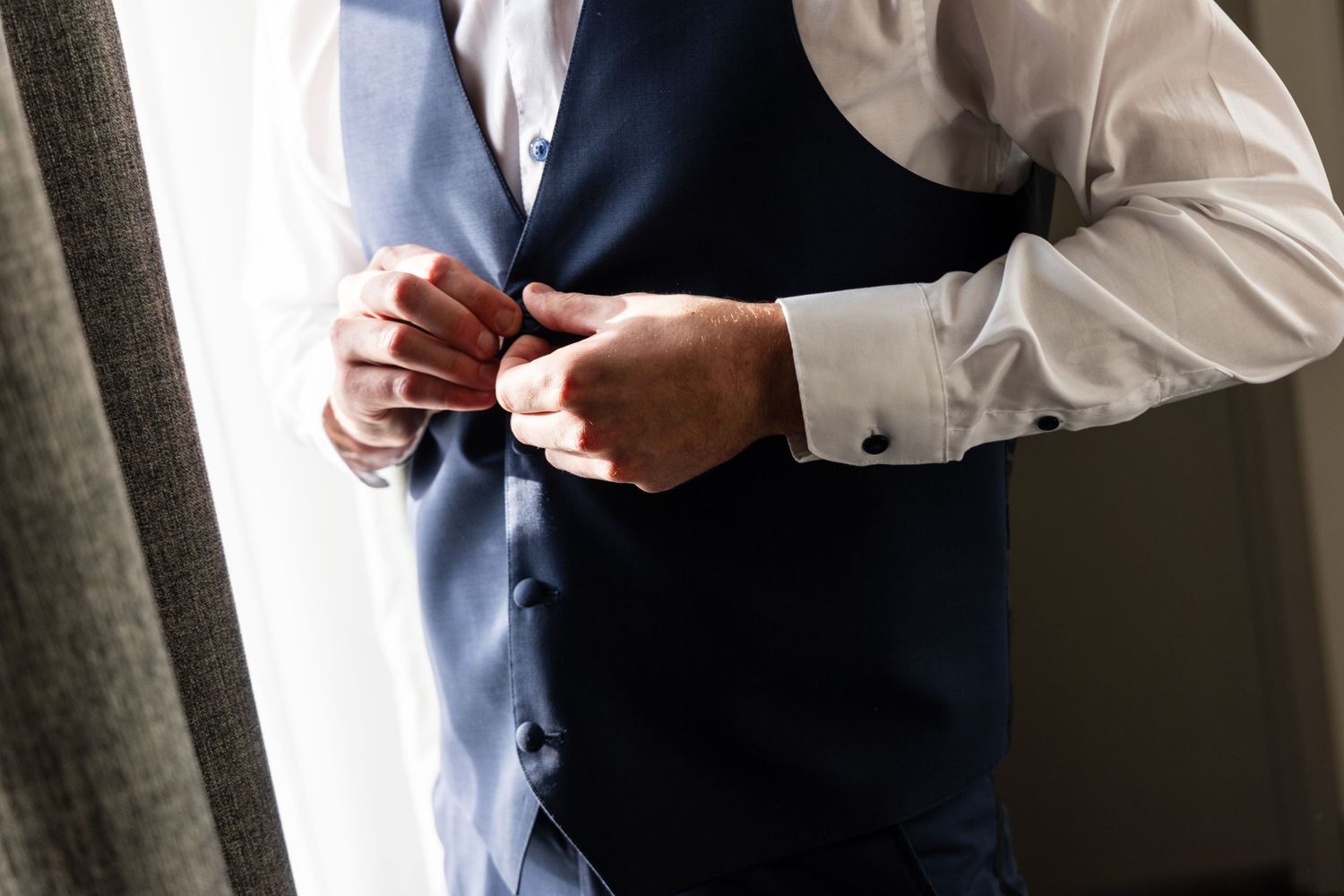 Person adjusting formal wear buttons on a navy blue vest near a window.