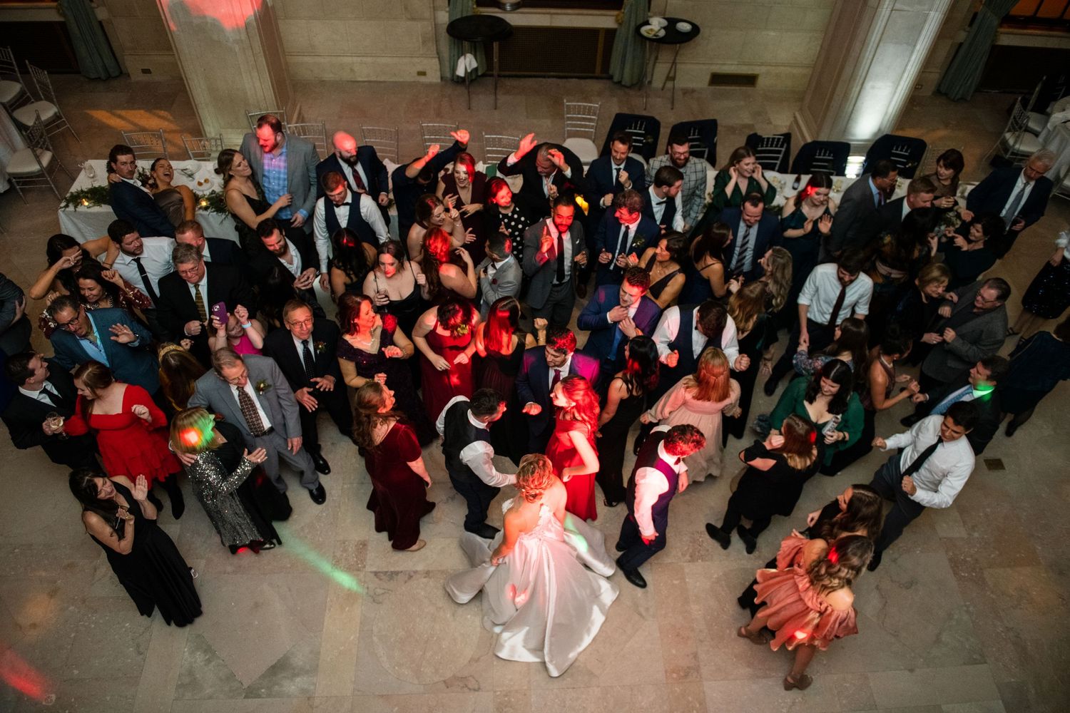 A large group of wedding guests dancing together on the dance floor under red lighting.