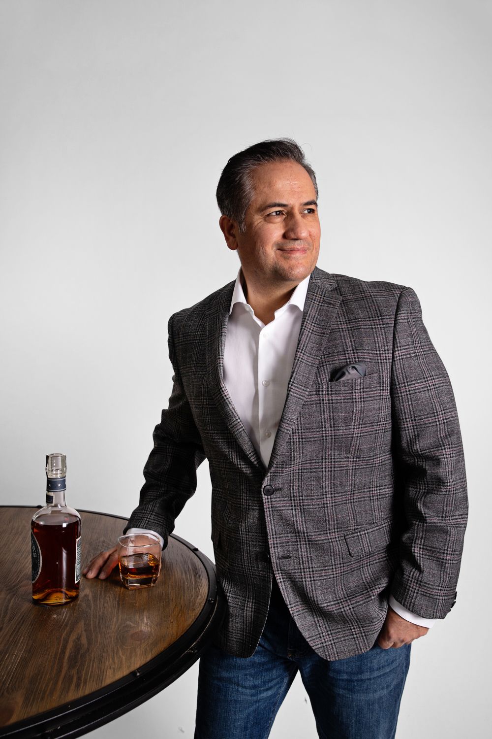 Sophisticated man in plaid blazer standing by wooden table with whiskey glass.