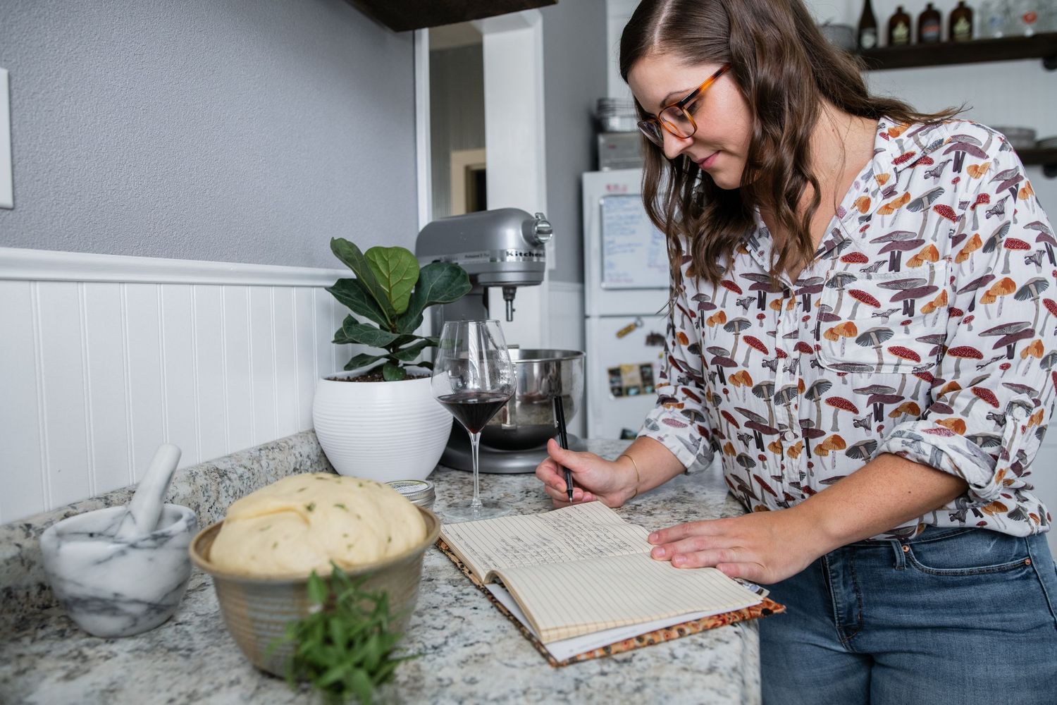 A cookbook being read on a marble counter with bread dough and kitchen ingredients nearby.