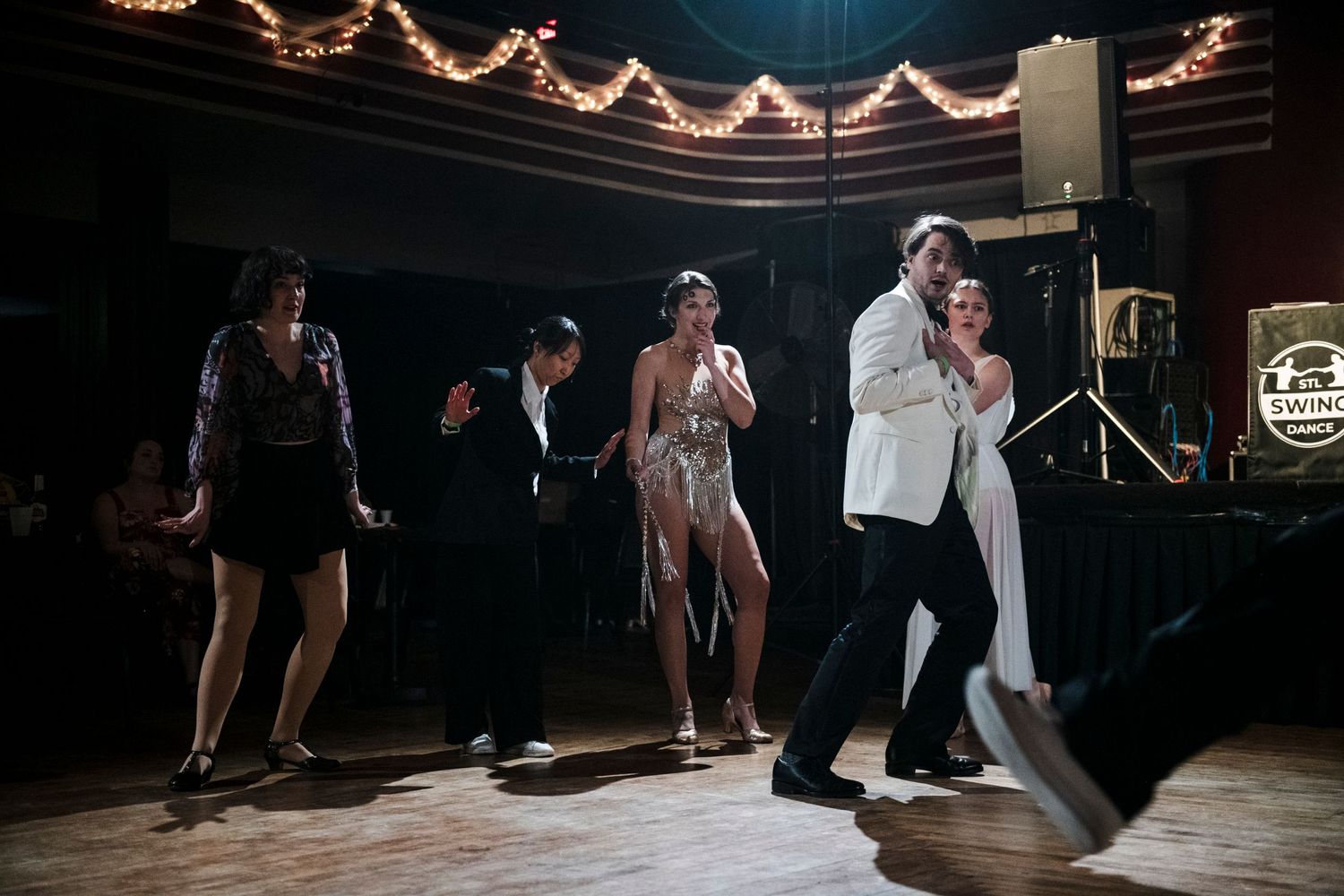 Dancers perform energetic moves on a dimly lit stage with string lights and vintage decor.